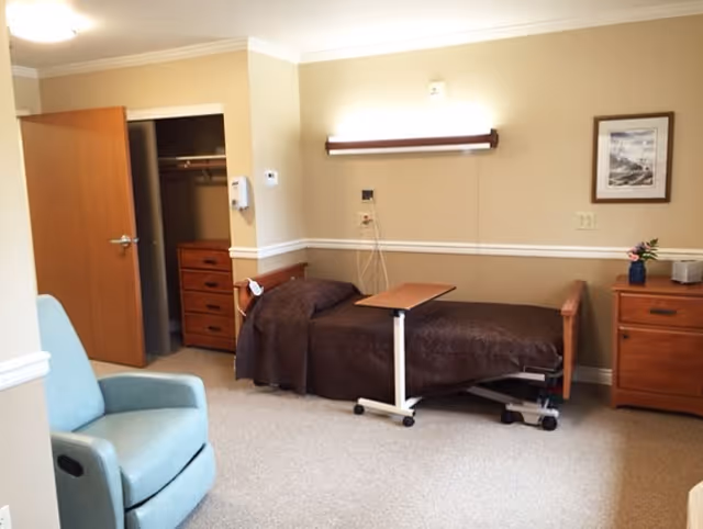 Furnished senior living bedroom with a single hospital-style bed, overbed table, recliner, and dressers in neutral decor.