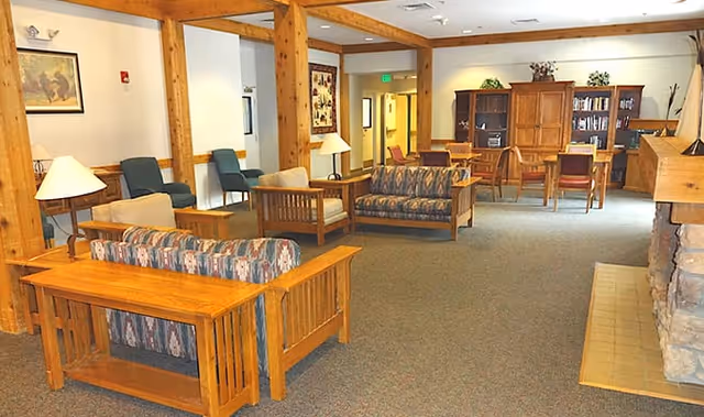 A spacious living room area in an assisted living facility featuring wooden framed sofas with patterned cushions, wooden side tables with lamps, a stone fireplace on the right, and a wooden cabinet with books and decorative plants in the background. The room has carpeted flooring and wooden support beams.