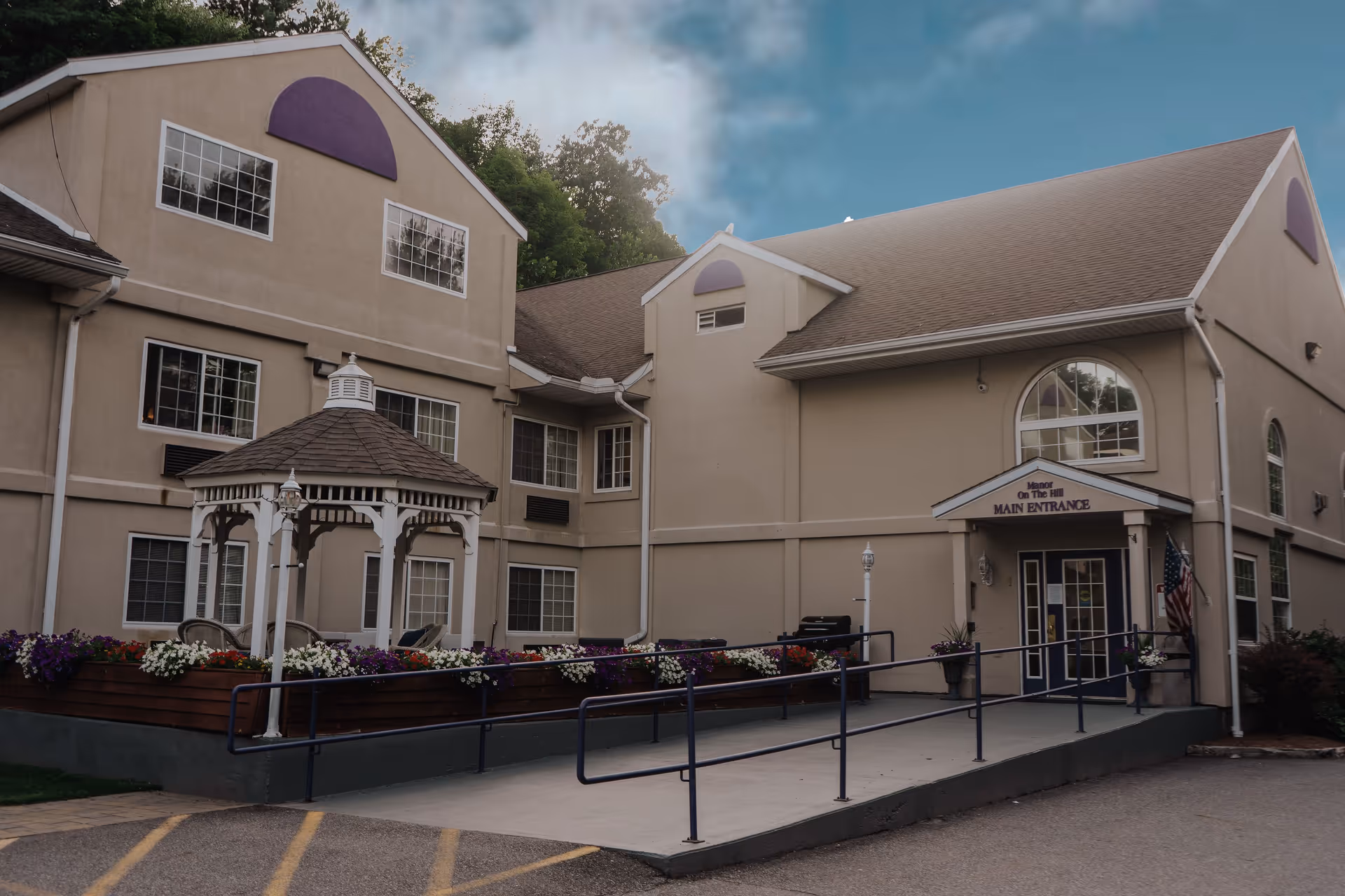 Exterior view of Manor On the Hill senior living facility showing the main entrance with a ramp, a small white gazebo surrounded by flower beds, and a beige building with multiple windows under a cloudy sky.