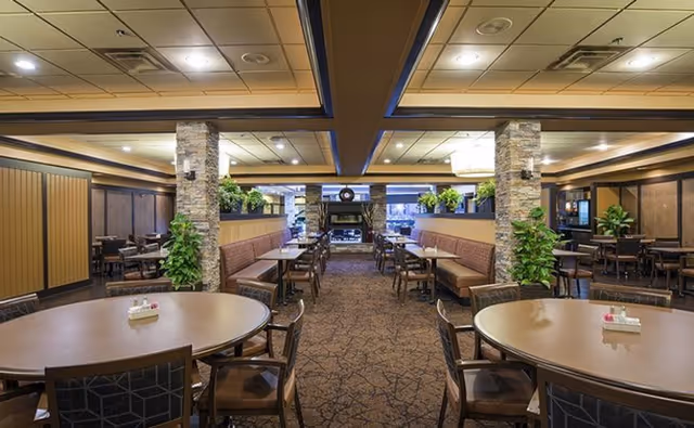 Interior view of a senior living community dining room with round and rectangular tables, chairs, cushioned bench seating along stone pillars, potted plants, and a fireplace in the background.
