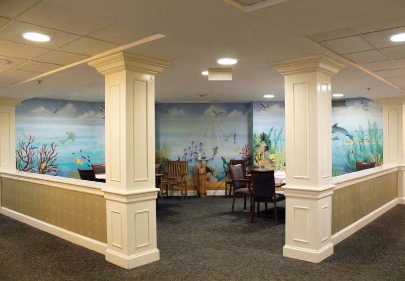 Interior common area with several wooden chairs and tables arranged around a corner. The walls feature a colorful underwater mural with fish, coral, and sea plants. The space is framed by white pillars and half walls with beige paneling.
