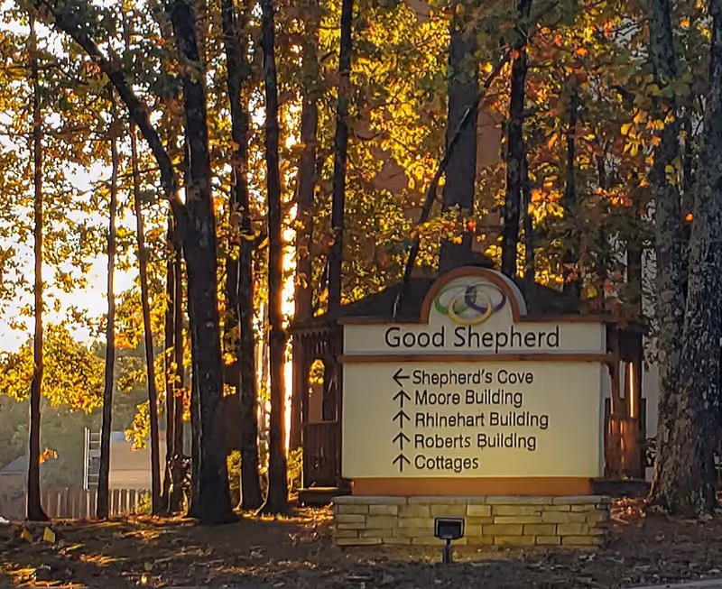 A wooden sign for Good Shepherd Community is displayed outdoors among tall trees with sunlight filtering through. The sign lists directions to Shepherd's Cove, Moore Building, Rhinehart Building, Roberts Building, and Cottages.