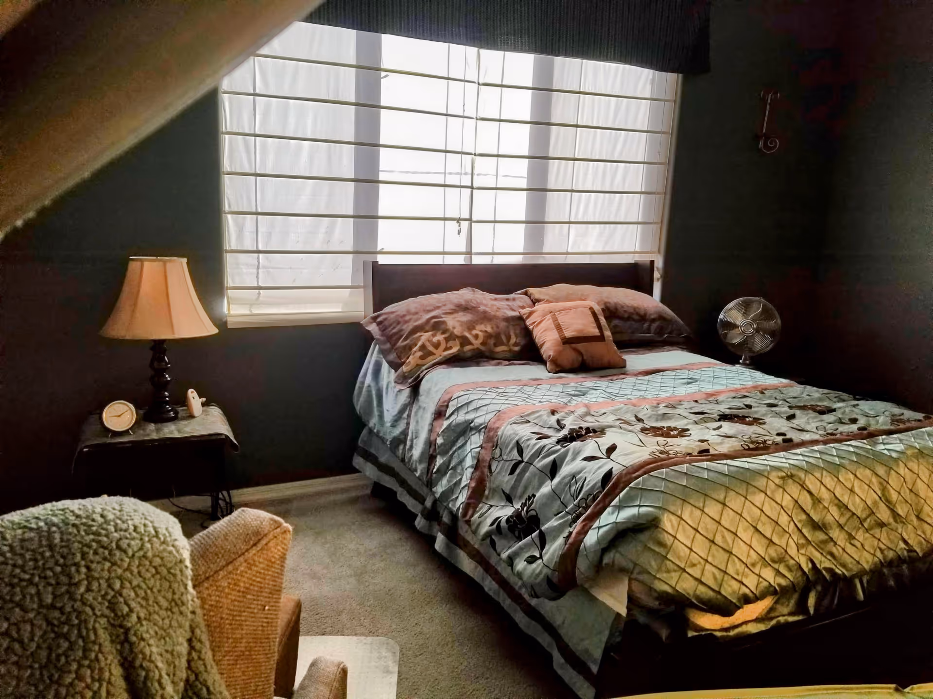 A cozy bedroom with a bed covered in a floral and quilted bedspread, several pillows, a small bedside table with a lamp and clock, a window with blinds letting in natural light, and a small fan on the floor near the bed.