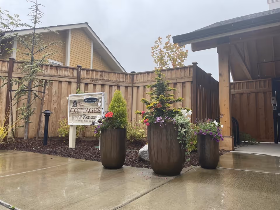 Outdoor entrance area of The Cottages of Renton facility with three large planters containing various flowers and greenery, a wooden fence, and a covered walkway. A sign reading 'Welcome The Cottages of Renton' is visible near the fence.