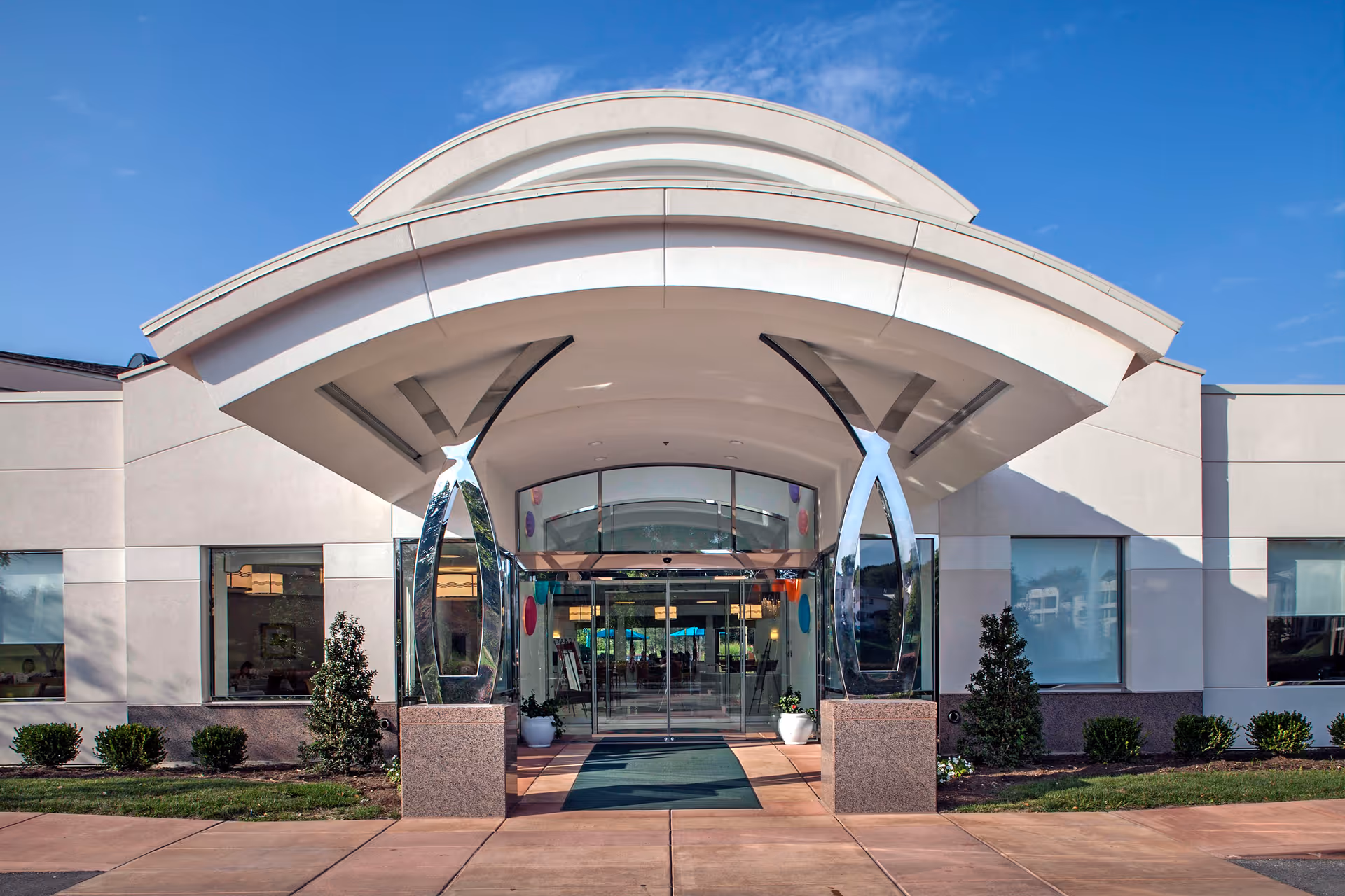 Front entrance of a modern senior living facility with an arched canopy, metal sculptures, glass doors, and landscaping.