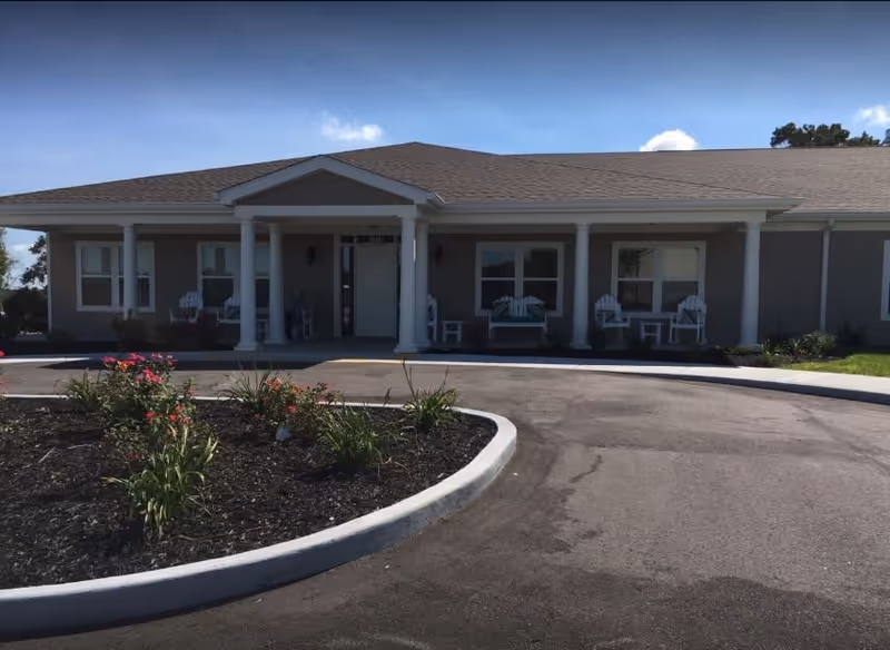 Front exterior view of a single-story senior living facility building with a covered entrance supported by white columns. There are several white chairs on the porch, and a landscaped area with plants and flowers is visible in the foreground under a clear blue sky.