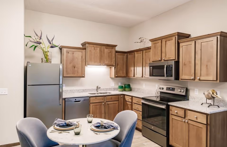 A modern kitchen with wooden cabinets, a stainless steel refrigerator, dishwasher, oven, and microwave. A small round dining table with two place settings and blue chairs is in the foreground. Decorative items include a vase with flowers on the refrigerator and two green vases on the countertop.