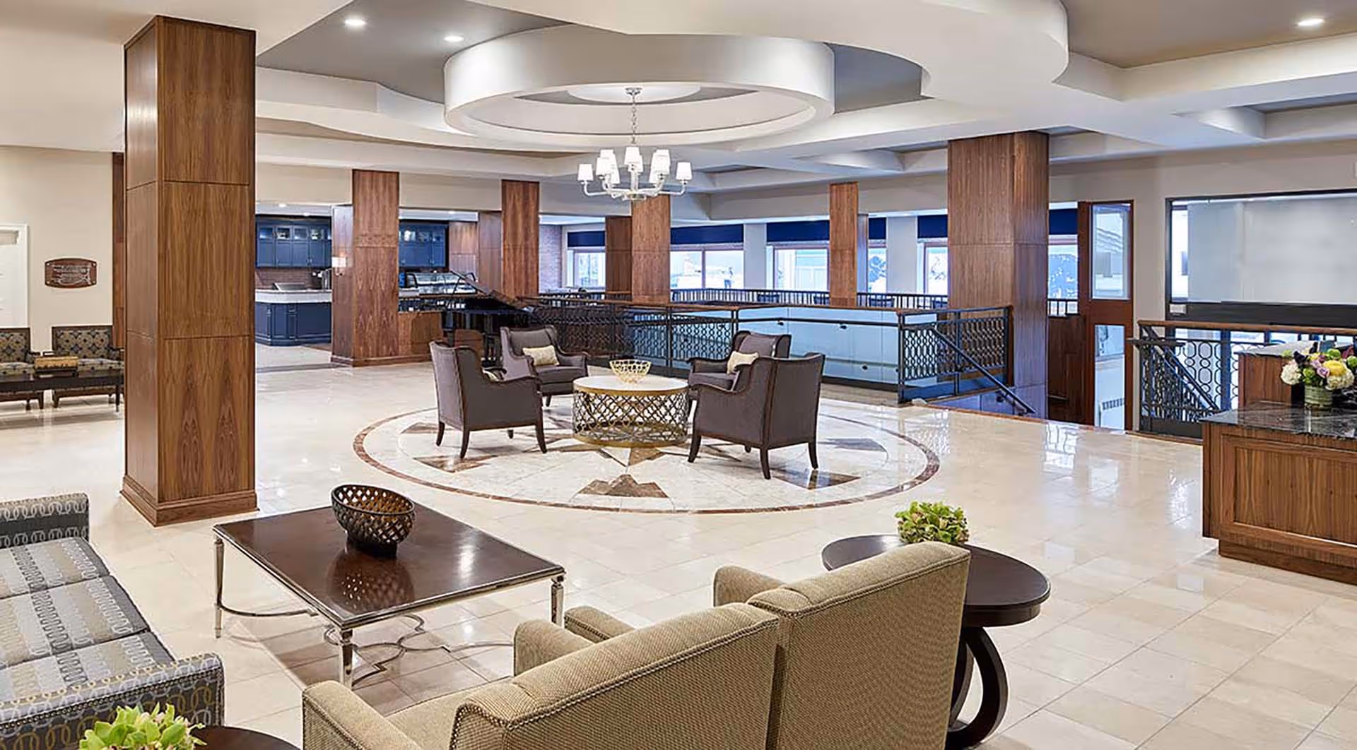 Spacious and well-lit common area in a senior living facility featuring comfortable seating arrangements with armchairs and sofas around coffee tables, wooden columns, a decorative ceiling light fixture, and large windows allowing natural light.