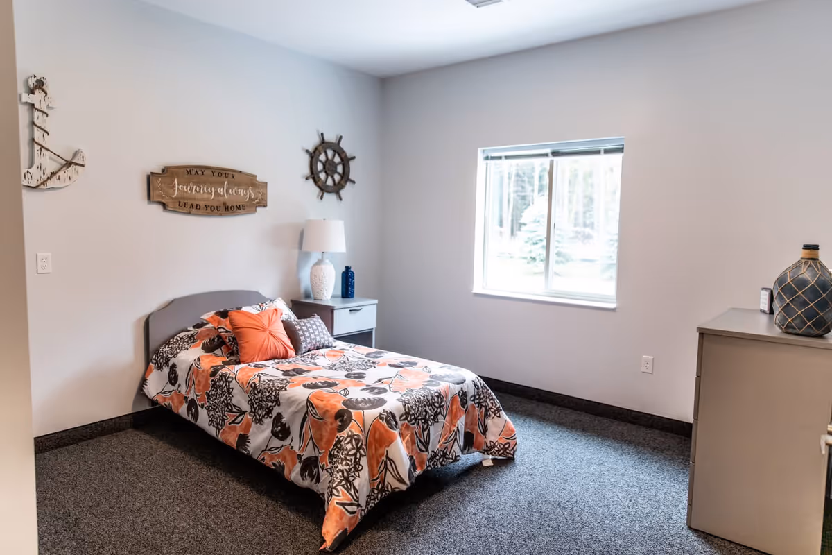 A simple bedroom with a single bed covered in a floral-patterned orange, black, and white bedspread. There is a small nightstand with a white lamp and decorative items next to the bed. The walls are light-colored with nautical-themed decorations including a wooden anchor, a ship's wheel, and a sign that reads 'May your journey always lead you home.' A window lets in natural light, and a dresser with a decorative vase is on the right side of the room.