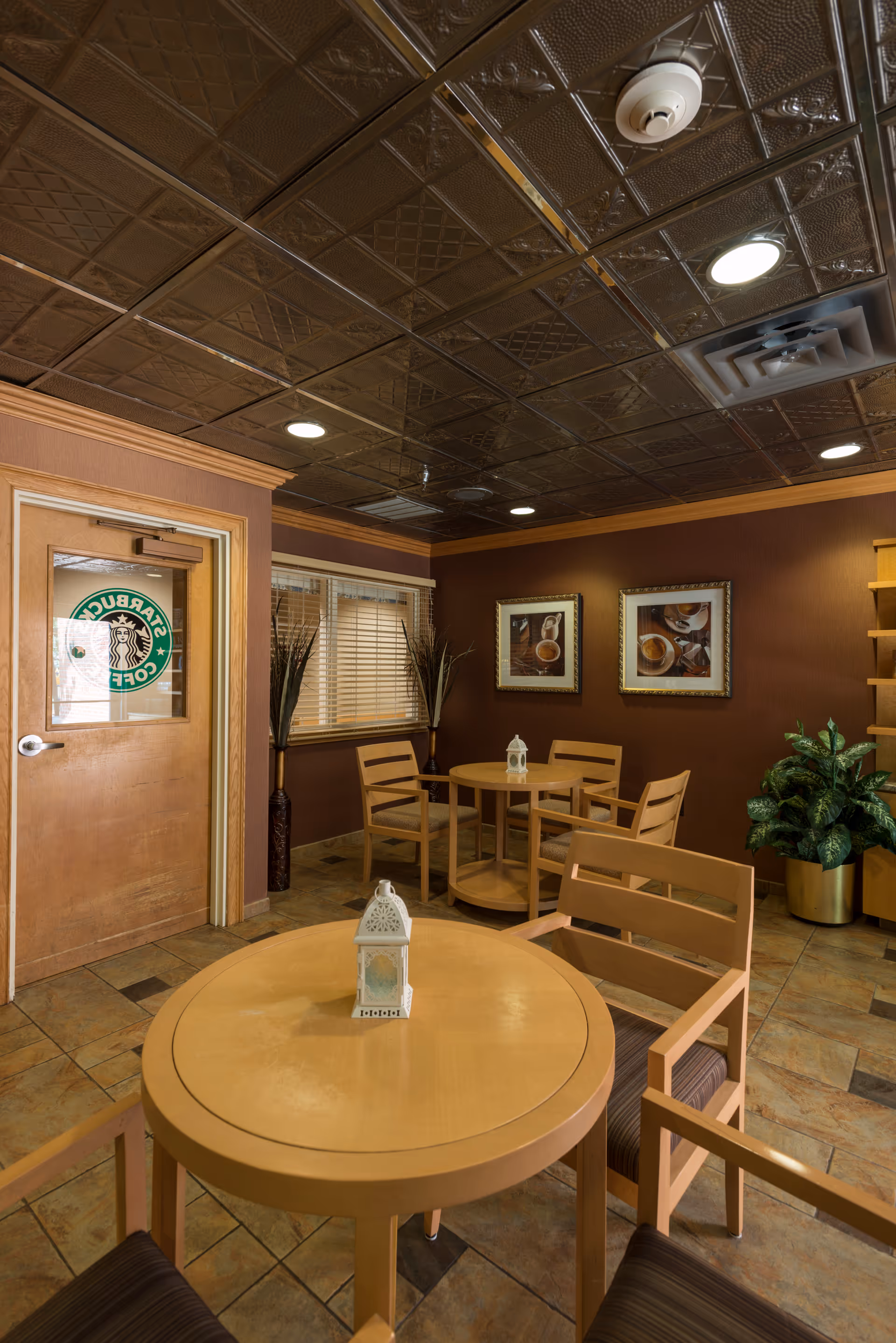 Interior view of a small seating area with round wooden tables and chairs. The room has a decorative ceiling with recessed lighting, brown walls adorned with framed coffee-themed pictures, and a door with a Starbucks Coffee logo on the glass. There are potted plants and window blinds in the background.