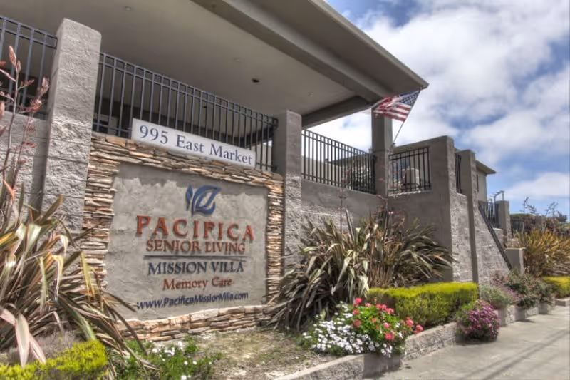 Exterior view of Pacifica Senior Living Mission Villa Memory Care facility with a stone and concrete sign displaying the address 995 East Marker. The building has a fenced balcony and an American flag is visible. There are plants and flowers along the sidewalk in front of the sign.