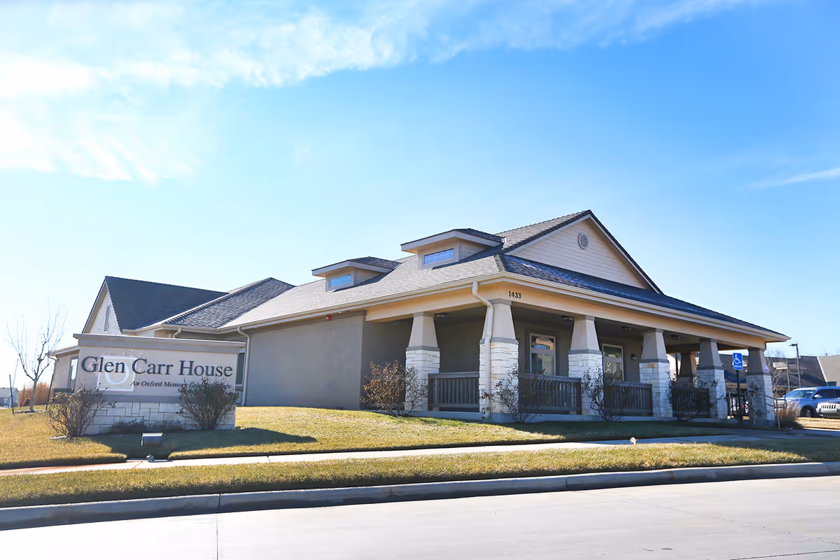 Exterior view of Glen Carr House Memory Care facility showing a single-story building with a covered porch, stone pillars, and a sign on the lawn with the facility name. The sky is clear and blue.