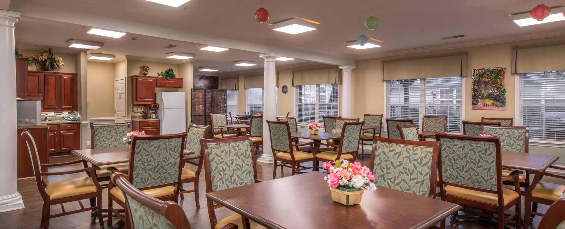 A well-lit dining room with multiple wooden tables and chairs featuring patterned upholstery. Each table has a small floral centerpiece. The room has large windows with blinds, beige walls, white columns, and a kitchen area with wooden cabinets and a white refrigerator in the background. Decorative hanging lanterns are visible on the ceiling.