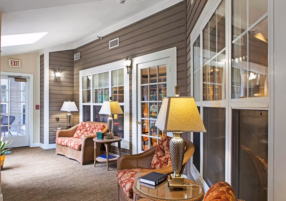 A cozy indoor seating area with wicker chairs and a loveseat featuring red floral cushions. There are two side tables with lamps and books on them. The walls are brown with white trim, and there are large windows and a glass door leading to another room. An exit door is visible at the end of the hallway.