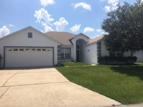 Front exterior of a single-story house with an attached two-car garage, driveway, lawn and a tree under a blue sky.