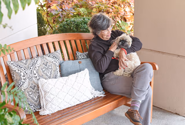 An elderly woman sitting on a wooden bench outdoors, holding and petting a small dog. The bench has decorative pillows, and there are plants and colorful foliage in the background.