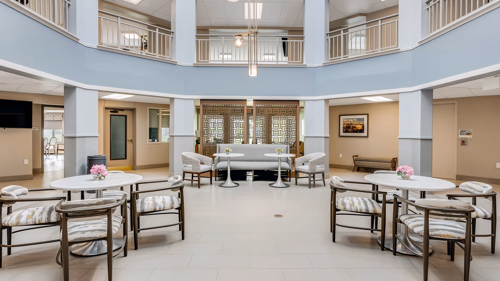 A spacious and bright common area in a senior living facility with round tables and cushioned chairs arranged neatly. The room features a two-story ceiling with a balcony railing on the upper level, light blue and beige walls, and decorative panels in the background. Small flower arrangements are placed on each table.
