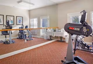 Exercise room with stationary bikes, large wall mirror, carpeted floor, and windows with blinds. A table with decorative items is visible near the windows.