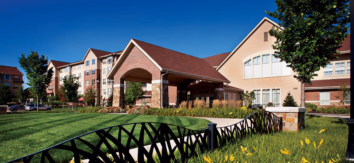 Exterior view of Tallgrass Creek Senior Living Community showing a large building with a covered entrance, well-maintained green lawn, trees, and a decorative black metal fence under a clear blue sky.