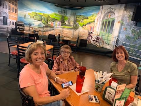 Three women sit at a dining table with drinks and menus in a communal dining area featuring a painted mural and small stage.