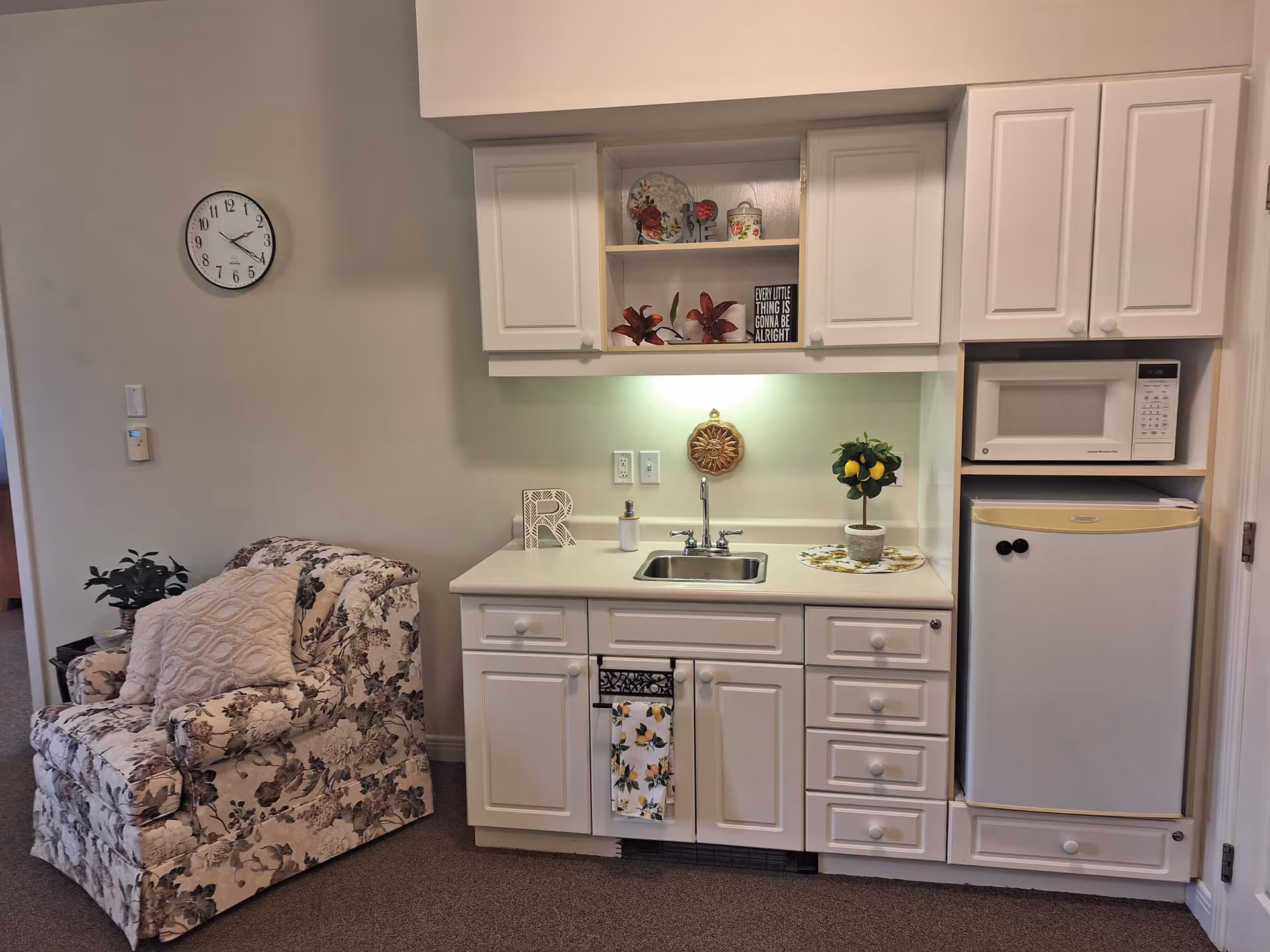 Small kitchenette with white cabinets, a sink, microwave and mini-fridge next to a floral upholstered armchair.