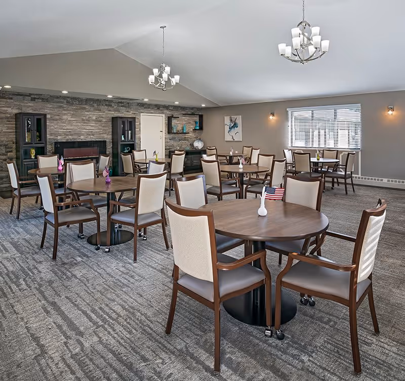 A spacious dining room with multiple round wooden tables and beige cushioned chairs. The room has a carpeted floor, a stone accent wall with built-in shelves and cabinets, and two modern chandeliers hanging from the ceiling. There is a window with blinds on the right wall and small decorative items on the tables, including a small American flag on one table.