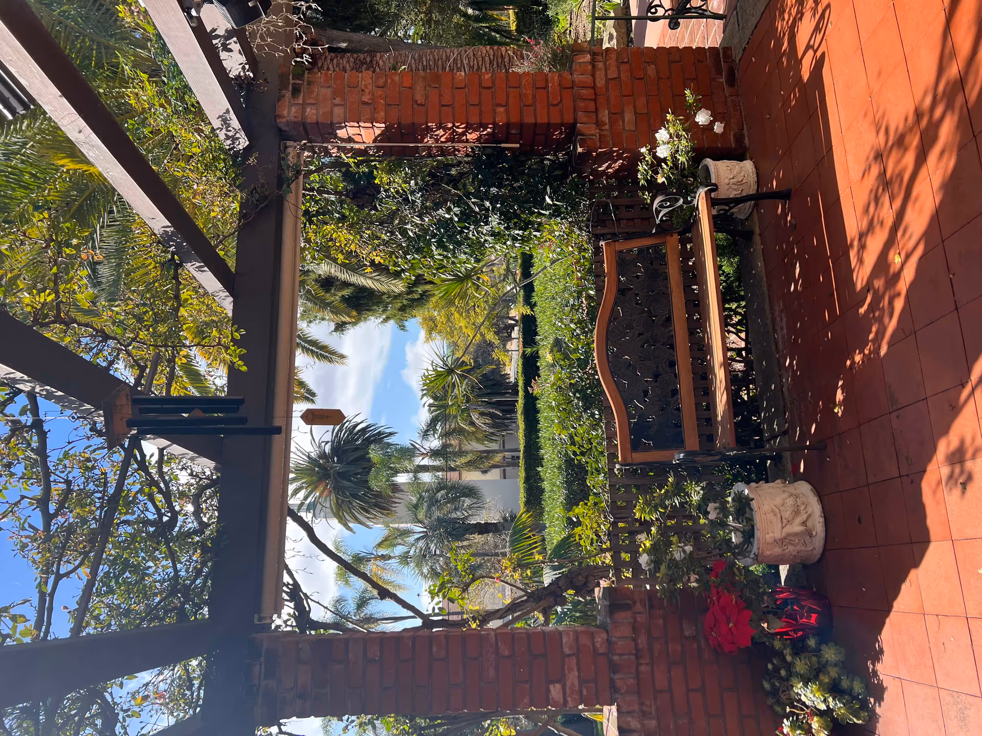 A sunny outdoor patio area with a decorative metal and wood bench, surrounded by potted plants and flowers. The patio has red tiled flooring and brick pillars supporting a wooden pergola with climbing plants. In the background, there are various green shrubs and palm trees under a blue sky with some clouds.