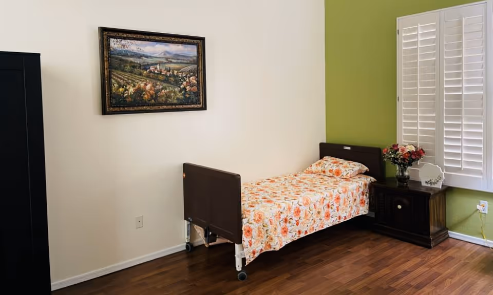 A small bedroom with a single bed covered in a floral bedspread. Next to the bed is a wooden nightstand with a vase of flowers and a decorative plate. The wall behind the bed is painted green, and the adjacent wall is white with a framed landscape painting. There are white plantation shutters on the window, and the floor is wooden.