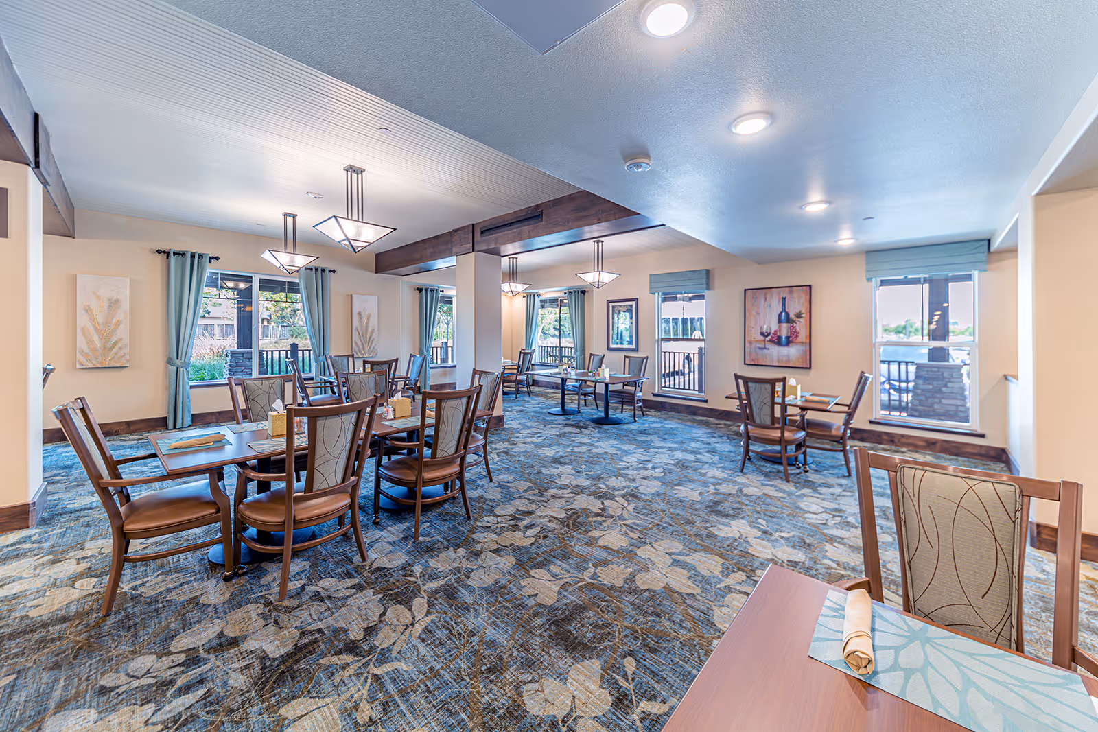 Spacious dining room with multiple wooden tables and chairs, patterned carpet, pendant lighting, and windows with blue curtains.