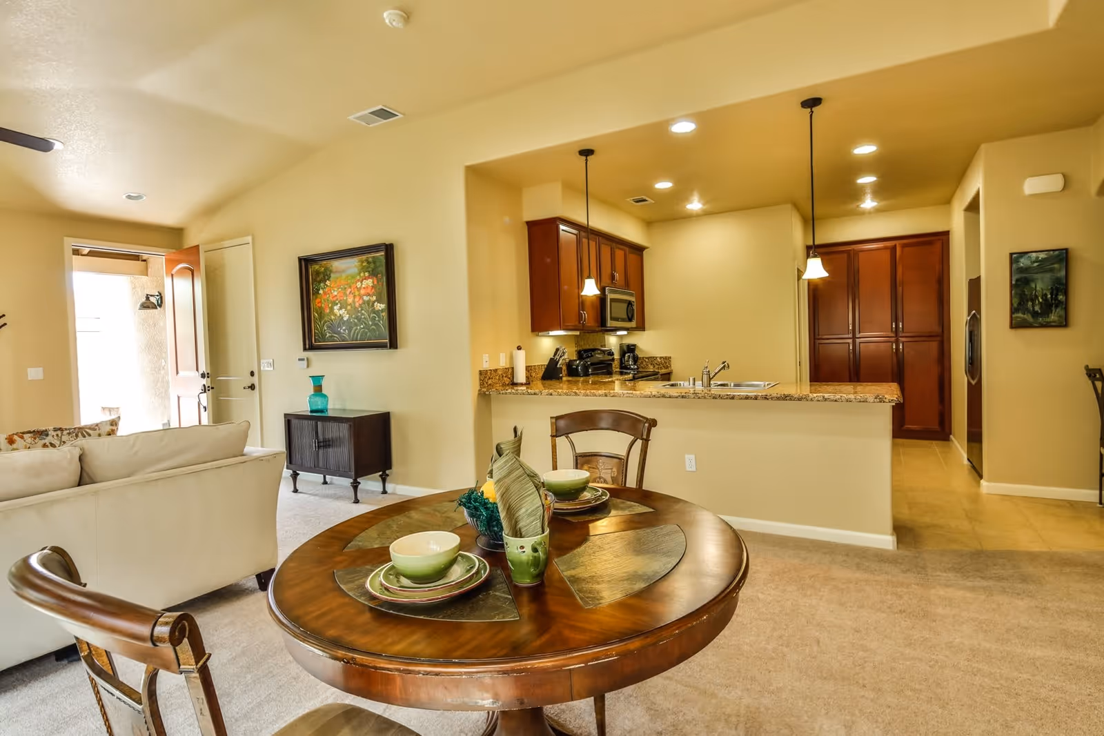 Interior view of a senior living facility showing a cozy living room with a beige sofa, a small dark wooden cabinet with a turquoise vase, and a round wooden dining table set with green dishes. The kitchen area features granite countertops, wooden cabinets, stainless steel appliances, and pendant lights hanging above the counter.