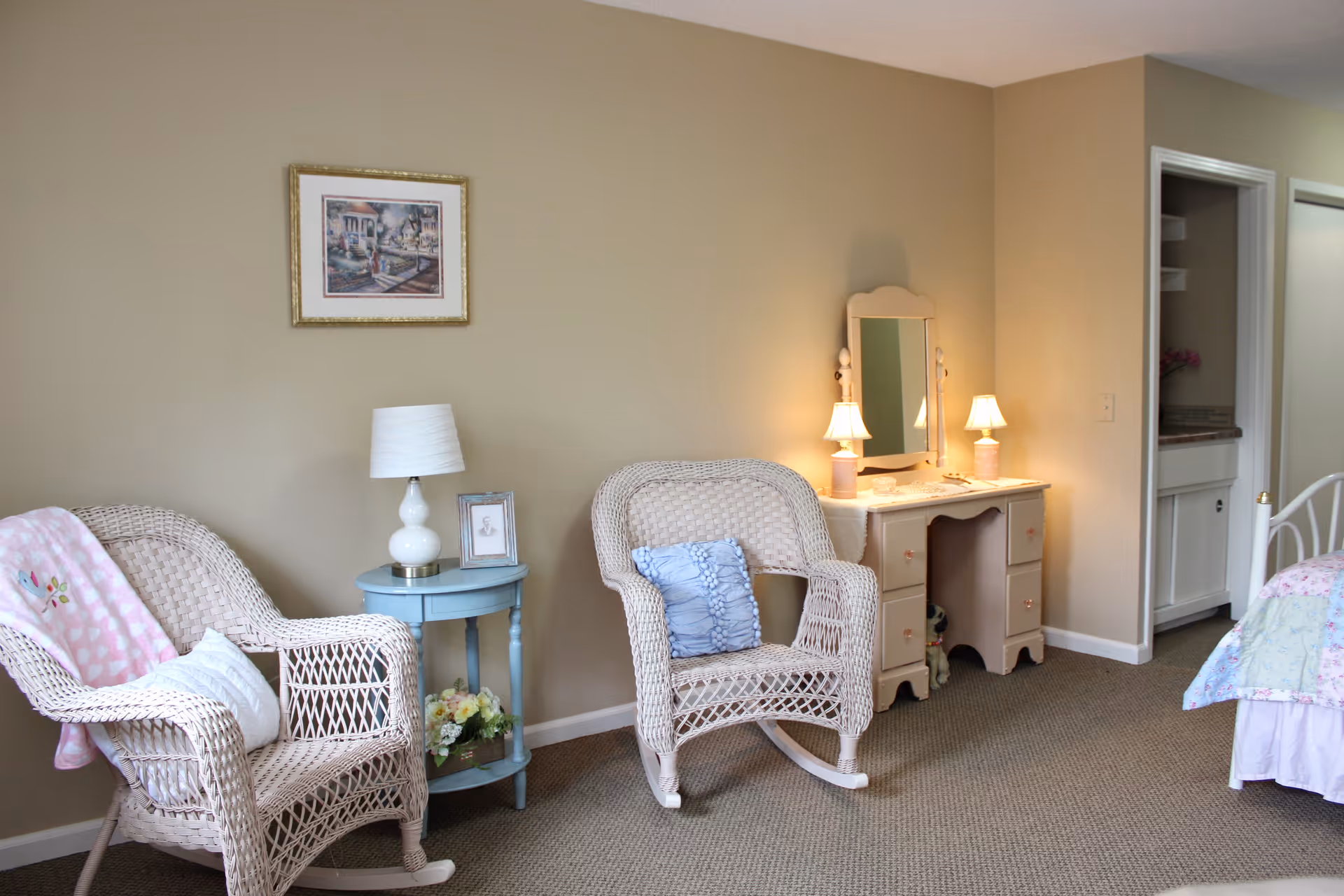 A cozy senior living room area with two wicker rocking chairs, one with a pink blanket and white pillow, and the other with a blue pillow. Between the chairs is a small round blue table with a white lamp and a framed photo. On the right side, there is a white vanity desk with a mirror and two small lamps, and part of a bed with a floral quilt is visible. The walls are beige, and the floor is carpeted.