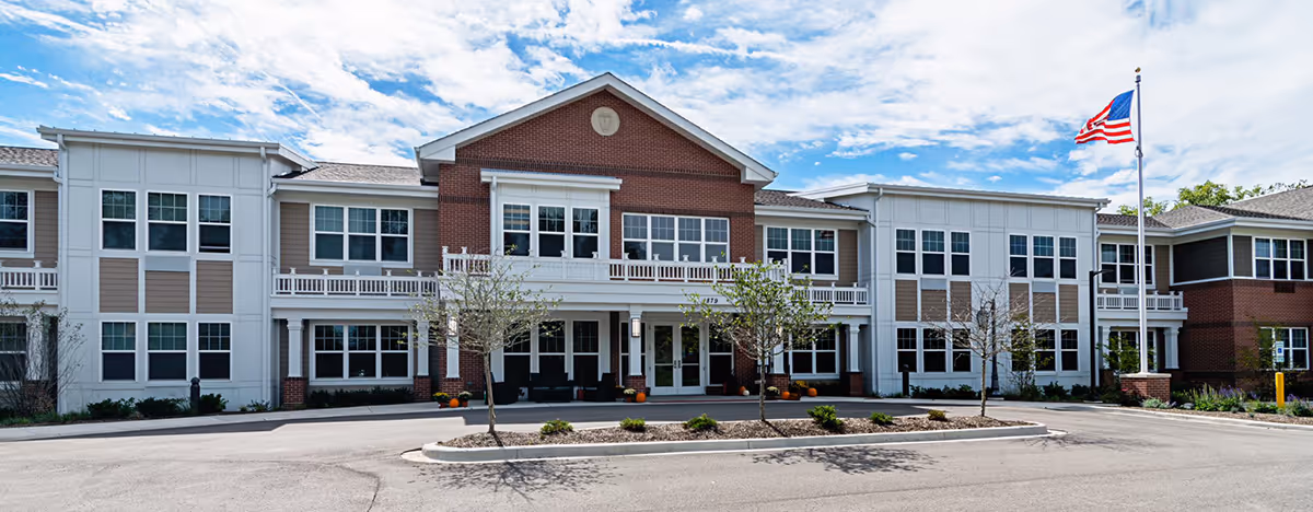 Front exterior view of a two-story senior living facility building with a brick and white facade, multiple windows, a covered entrance, small trees, and an American flag on a flagpole against a partly cloudy sky.