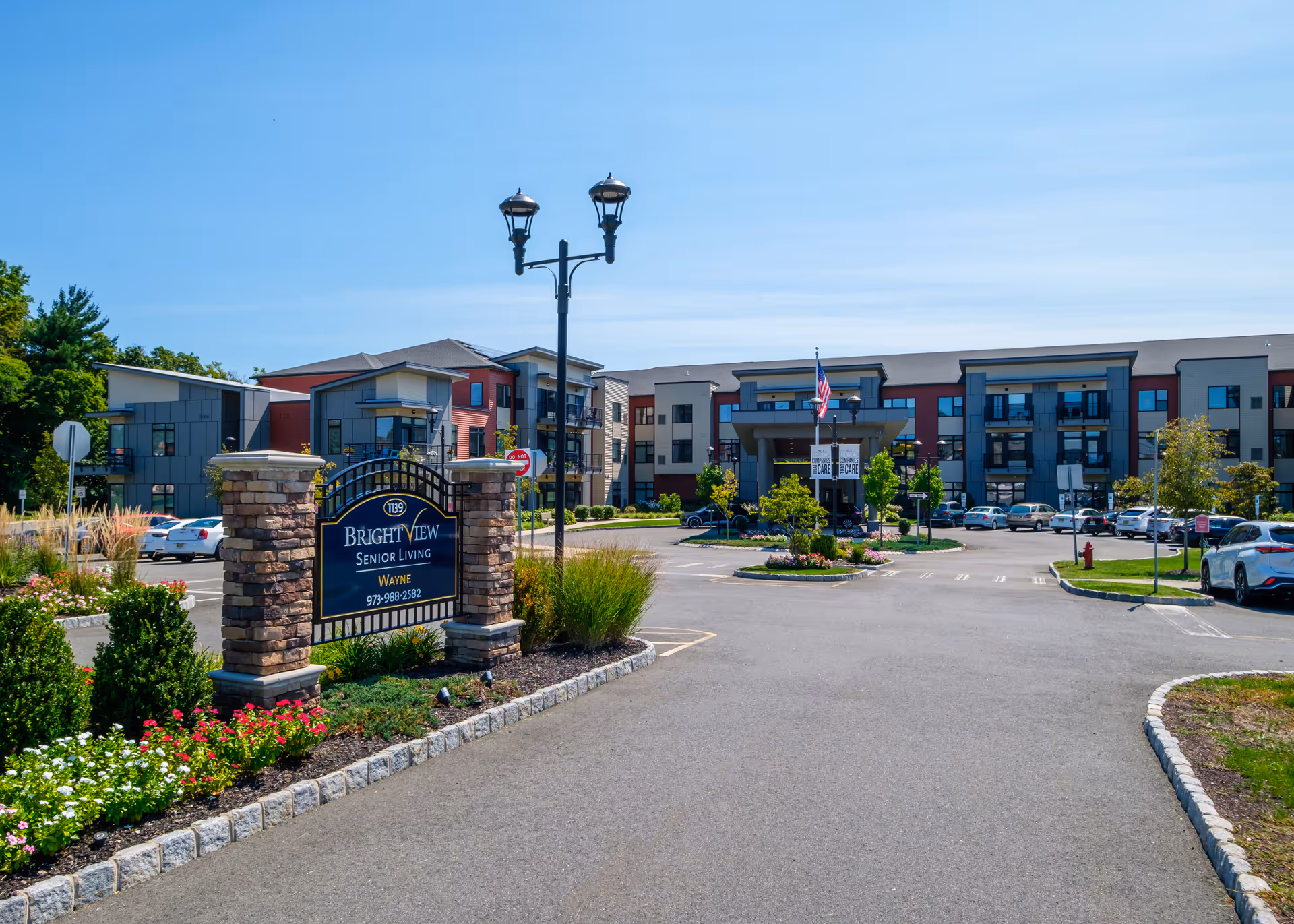 Exterior view of Brightview Wayne senior living facility showing a large parking lot, landscaped flower beds, and a multi-story building with balconies under a clear blue sky.