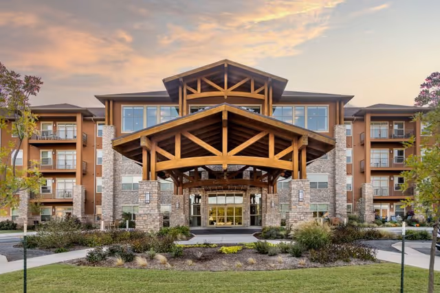 Front exterior view of Legacy Village of Stillwater, a multi-story senior living facility with a large wooden entrance canopy, stone pillars, and landscaped garden in front under a partly cloudy sky at sunset.