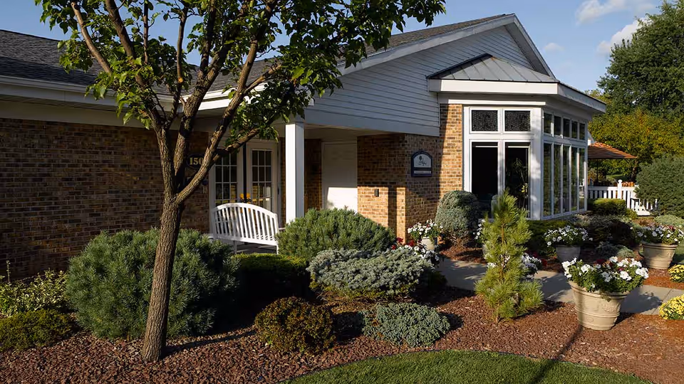 Exterior view of a senior living facility building with brick and white siding, surrounded by well-maintained landscaping including bushes, small trees, and flower pots. A white bench is visible near the entrance under a covered porch.