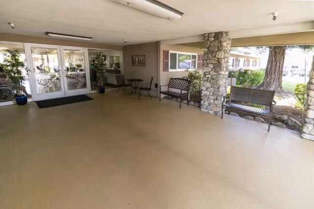 Covered outdoor entrance area of Claremont Care Center with stone pillars, metal benches, small tables with chairs, and glass double doors leading inside.