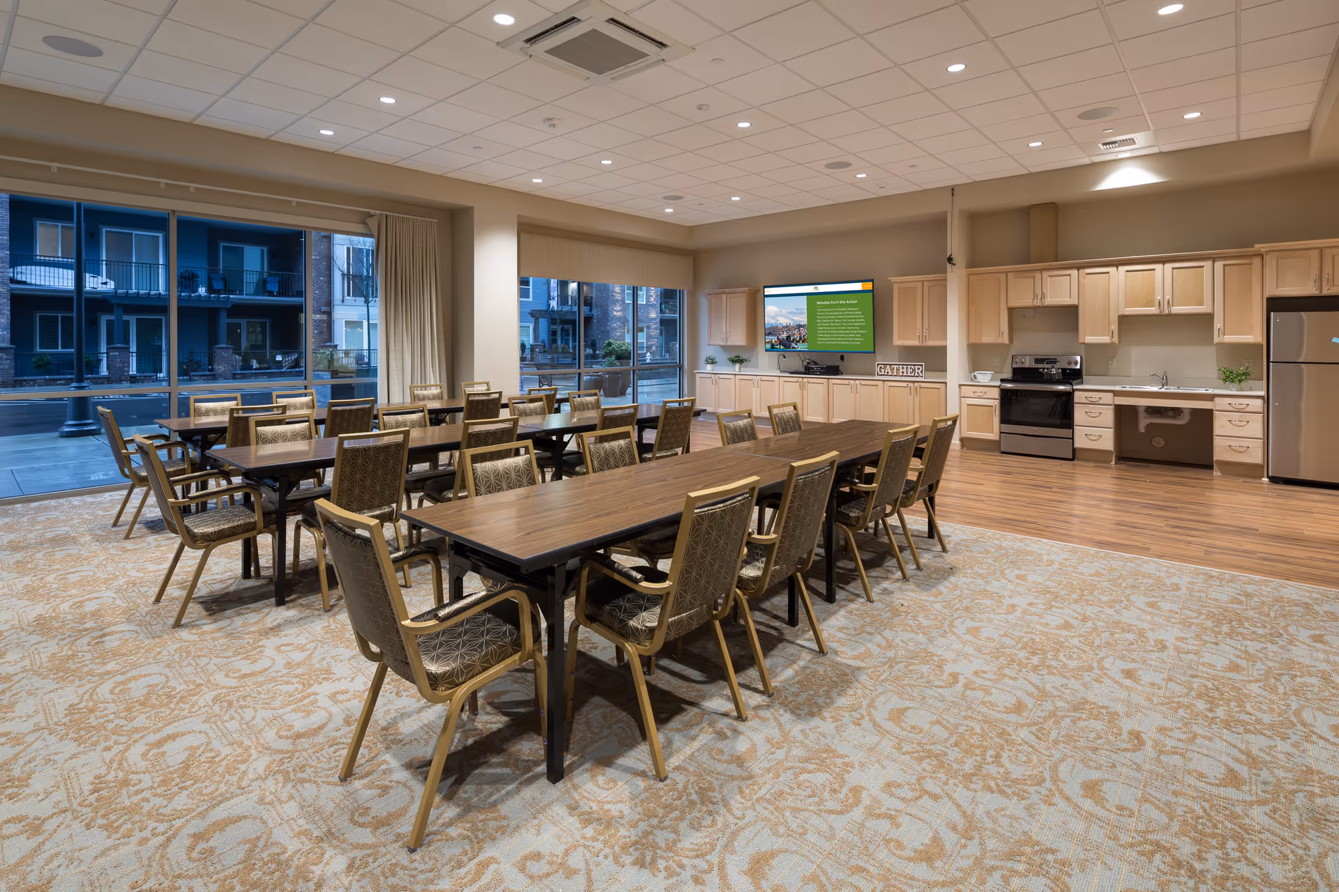 A spacious dining area with multiple wooden tables and cushioned chairs arranged neatly. The room features large windows with views of an outdoor area and adjacent buildings. The back wall has a kitchen setup with light wood cabinets, a stove, dishwasher, refrigerator, and a large flat-screen TV mounted above the counter. The carpet has a decorative pattern, and the ceiling has recessed lighting.