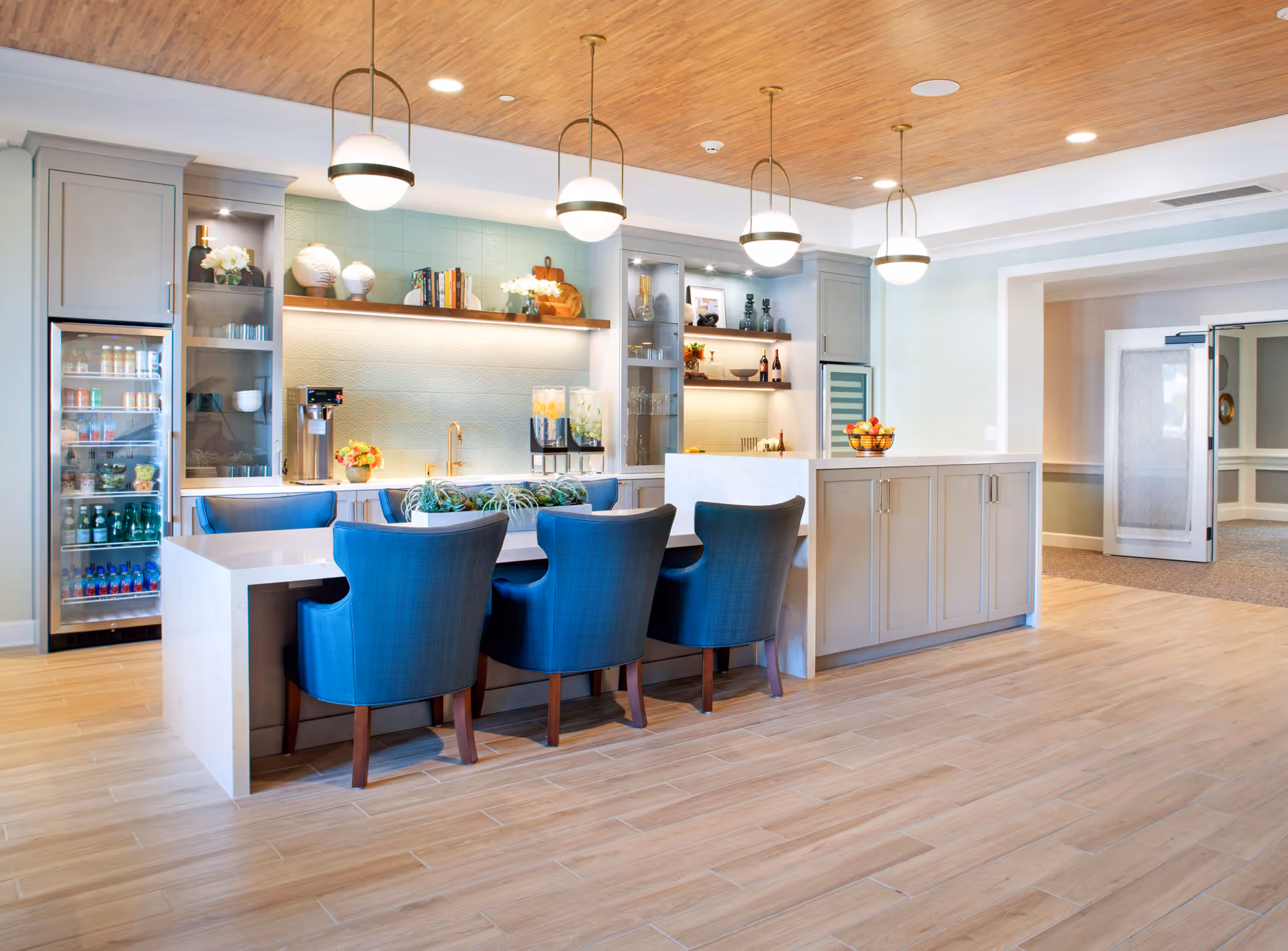 Bright communal kitchen and bar area with blue upholstered chairs, a marble island, pendant lights, and a stocked beverage refrigerator.