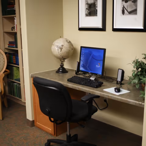 Small workstation with a desktop computer, keyboard and mouse, globe, office chair, and framed photos on a beige wall.