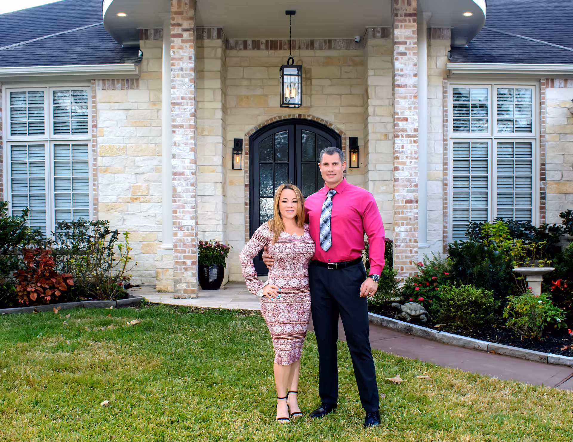 A man and a woman standing on a green lawn in front of a stone house with a black front door and large windows with white shutters. The woman is wearing a patterned dress and black heels, and the man is wearing a bright pink shirt with a patterned tie and black pants. There are plants and shrubs along the walkway leading to the entrance.
