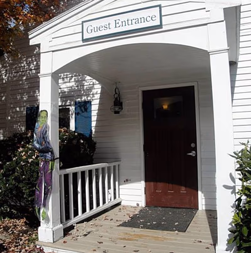 Covered porch entrance with a wooden door and a sign above reading 'Guest Entrance'. The porch has white railings and siding, with bushes on either side and a Halloween decoration of a green-faced figure attached to the left porch post.