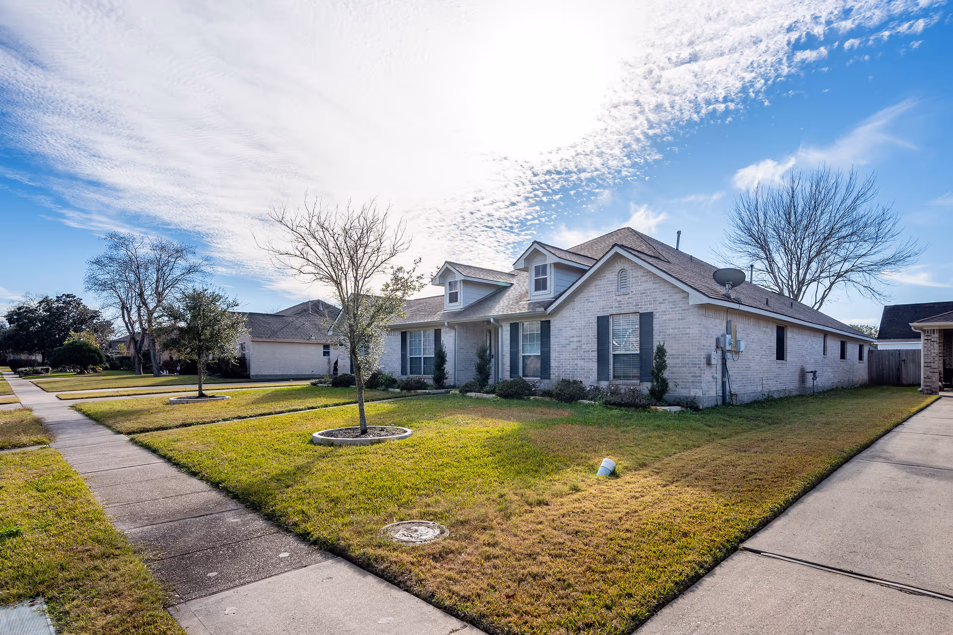 Single-story brick house with a manicured front lawn, small trees, driveway and sidewalk under a bright blue sky.