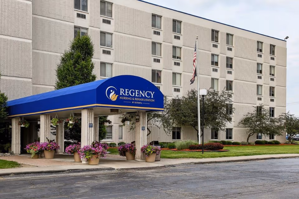 Exterior view of a multi-story nursing and rehabilitation facility with a blue awning entrance labeled 'Regency Nursing & Rehabilitation by Olympia'. The building is surrounded by green landscaping, potted flowers, and an American flag on a flagpole.