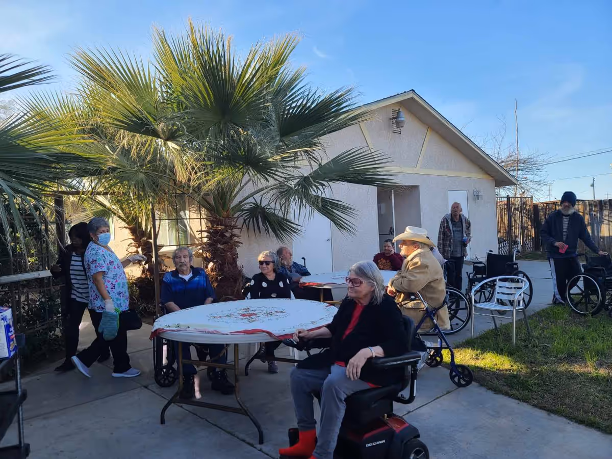 A group of residents and staff sit and stand around tables under palm trees in an outdoor courtyard next to a single-story building.