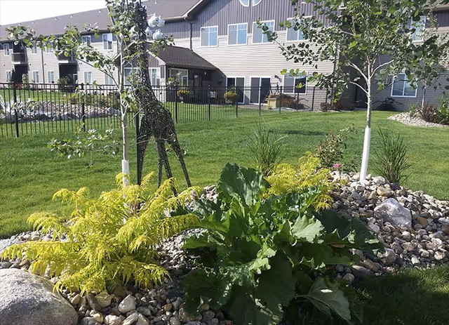 A landscaped outdoor area with green grass, small trees, shrubs, and rocks in the foreground. A wireframe giraffe sculpture is visible among the plants. In the background, there is a multi-story building with beige siding and multiple windows.
