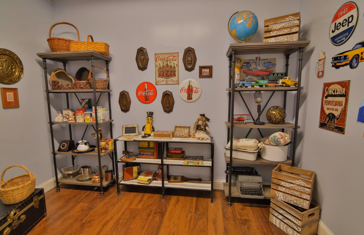 A room with wooden flooring and light gray walls featuring two tall metal shelves and one shorter metal shelf filled with various vintage items including baskets, old toys, books, a typewriter, and decorative objects. The walls are decorated with vintage-style signs and plaques, including Coca-Cola and Jeep logos.