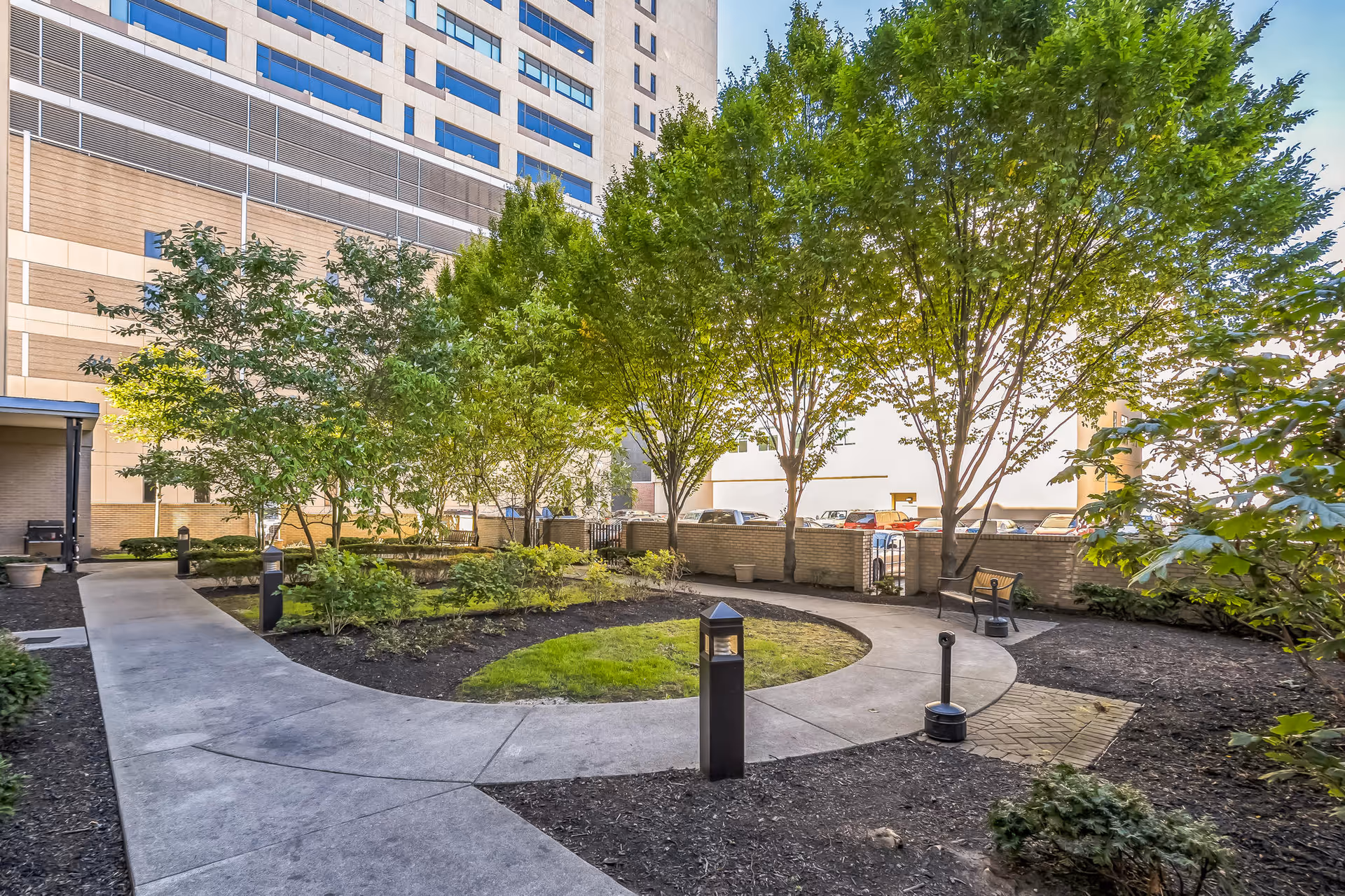 A peaceful outdoor courtyard area with a curved concrete walkway surrounding a small grassy patch. The courtyard is lined with green trees and shrubs, with a bench and lamp posts along the path. A tall building is visible in the background.