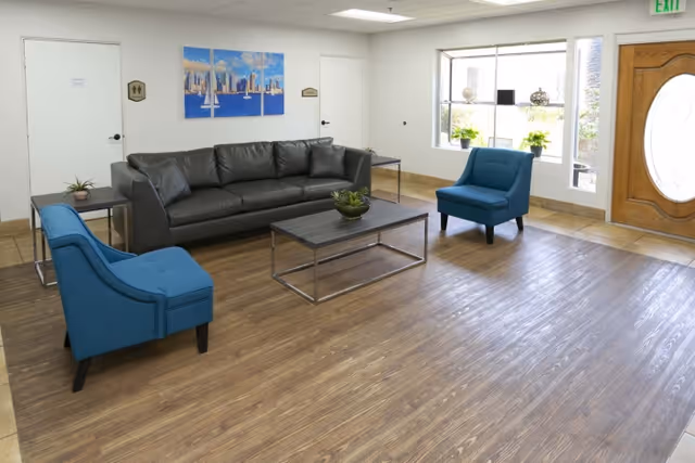 A bright and clean living room area with a dark gray leather sofa, two blue upholstered chairs, a wooden coffee table with a plant centerpiece, and a side table with a small plant. The room has wood-look flooring, white walls, a large window with decorative items on the sill, and a wooden door with an oval glass panel. There is a three-panel wall art piece depicting a city skyline above the sofa.