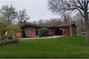 Single-story brick and wood building with a sloped roof, surrounded by green grass, trees, and a clear sky. There is a flagpole with an American flag near the building entrance.
