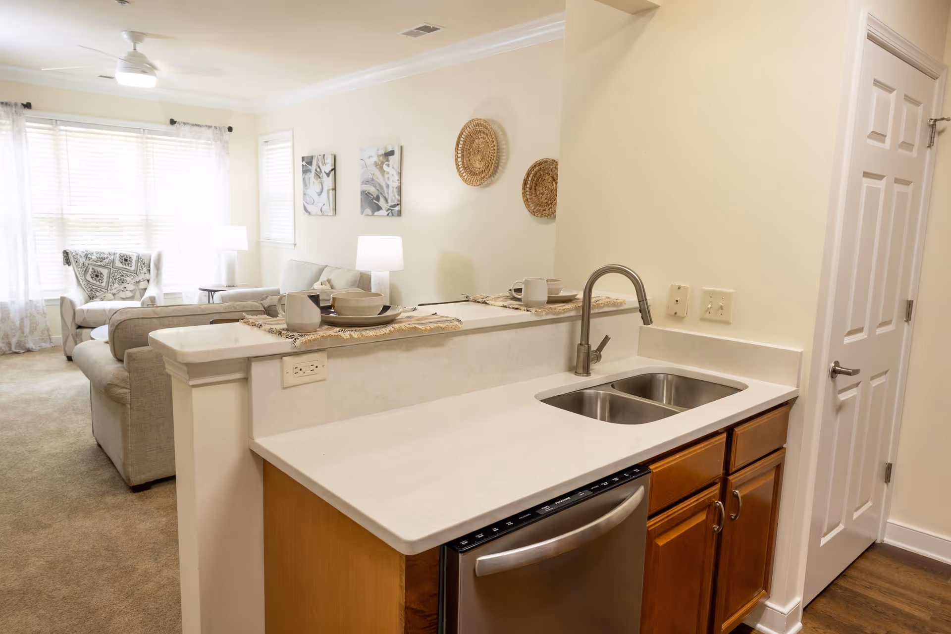 View of a modern kitchen area with a white countertop, double sink, and stainless steel dishwasher. The kitchen opens into a living room with beige sofas, a white armchair, two table lamps, wall art, and woven wall decorations. The room is well-lit with natural light coming through large windows with sheer curtains.